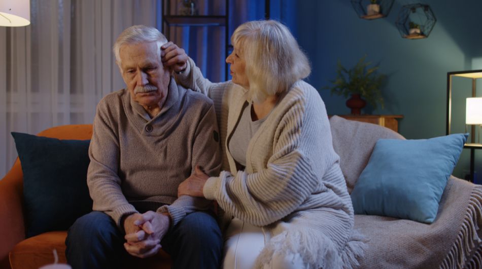 Älterer Mann sitzt mit verschlossenen Händen da und schaut traurig, während eine ältere Frau ihm tröstend über den Kopf streichelt.
