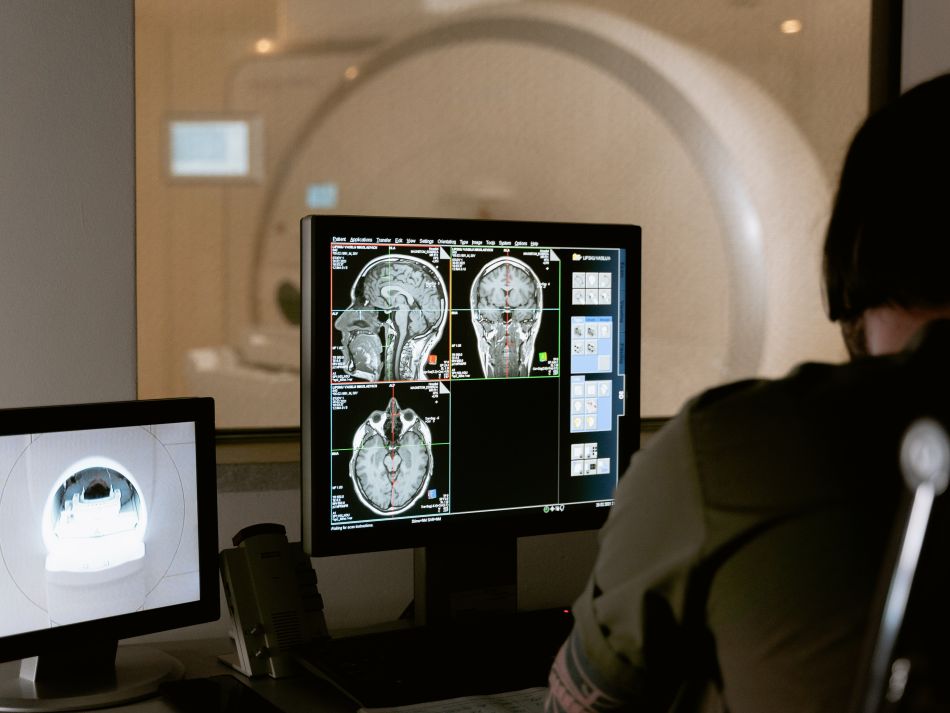 PC-Bildschirm mit MRT-Bildern des Gehirns, im Vordergrund ist eine Person zu sehen, die sich diese Bilder ansieht, im Hintergrund ein MRT-Gerät