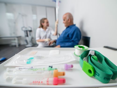 Arzthelferin und sitzender Patient im Hintergrund, im Bildvordergrund ein Tablett mit Probenröhrchen und Utensilien zur Blutabnahme.
