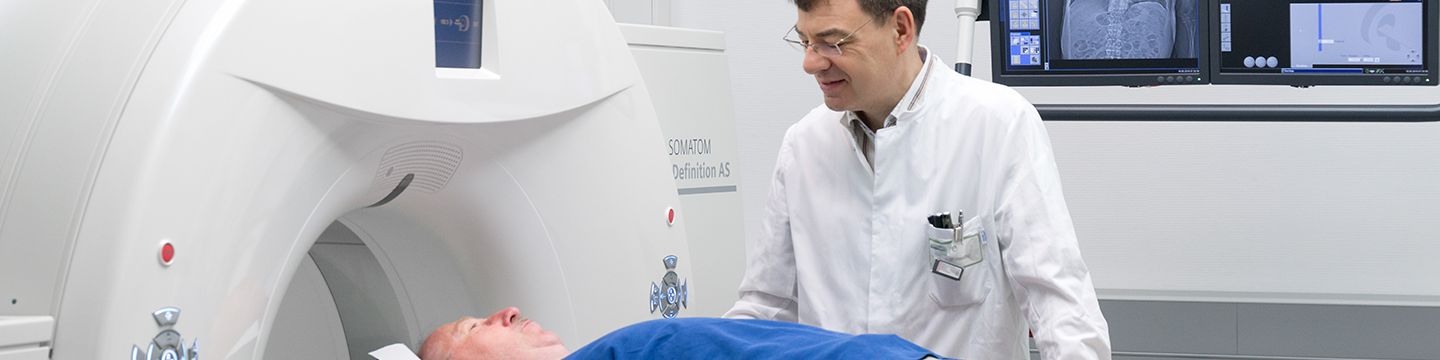   Ein Patient liegt auf der Liege eines Computertomographen, während der Arzt neben ihm steht und den Vorgang erklärt. Foto: Tobias Schwerdt © Krebsinformationsdienst, DKFZ