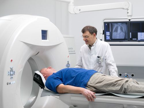  Ein Patient liegt auf der Liege eines Computertomographen, während der Arzt neben ihm steht und den Vorgang erklärt.