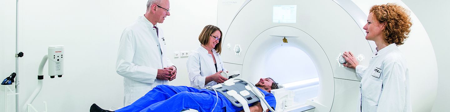 Ein Patient liegt auf der Liege eines Kernspintomographen, während Ärzte neben ihm stehen und den Vorgang erklären.