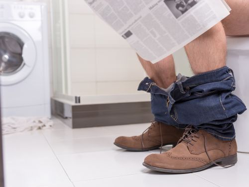 Mann sitzt auf einer Toilette und liest Zeitung.