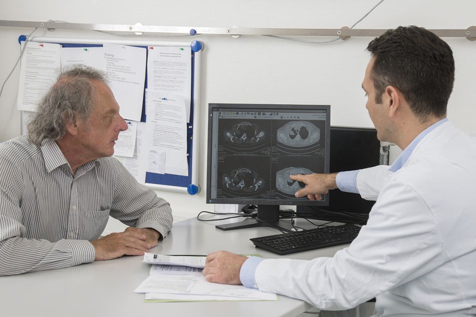 Lungenkrebspatient im Gespräch mit einem Arzt, der auf einen Montitor mit Röntgenaufnahmen der Lunge zeigt