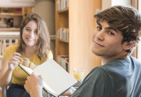 Zwei lächelnde Teenager: ein Junge und ein Mädchen. Der Junge hält ein Buch.