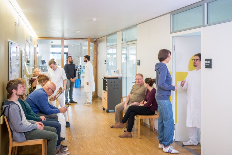 Mehrere Patienten und Patientinnen sitzen im Wartezimmer einer Tagesklinik oder Chemo-Ambulanz. Ärzte stehen im Flur und in der Tür zum Behandlungszimmer..