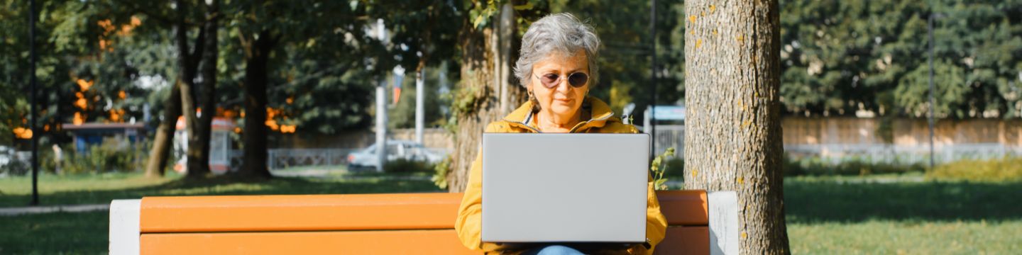 Frau mit Brille und lockeren, stilvollen Kleidung, die auf Laptop arbeiten, eine Nachricht schreiben, im Internet surfen, während sie im Freien auf der Parkbank sitzt. 