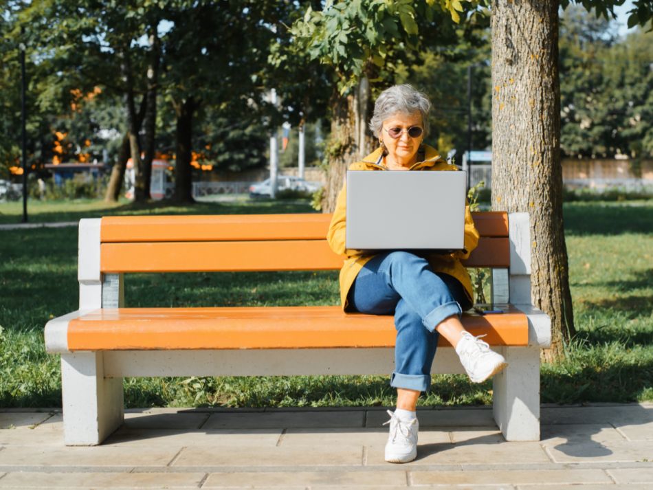 Frau mit Brille und lockeren, stilvollen Kleidung, die auf Laptop arbeiten, eine Nachricht schreiben, im Internet surfen, während sie im Freien auf der Parkbank sitzt. 