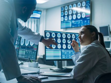 Eine Ärztin und ein Arzt diskutieren Befunde und Bilder eines Hirntumors, die auf einem PC-Bildschirm abgebildet sind. 