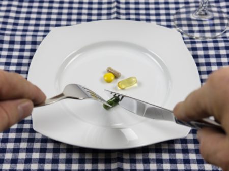 Nahrungsergänzungsmittel liegen auf einem weißen Teller, zwei Hände zerschneiden mit Gabel und Messer eine Kapsel.