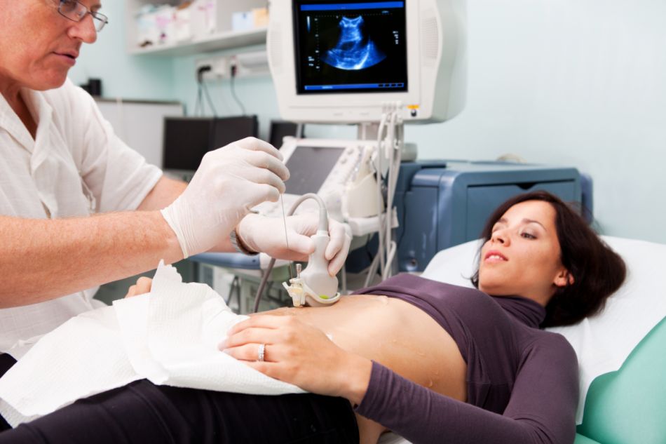 Eine junge Frau liegt auf einer Untersuchungsliege. Ein Arzt hält einen Ultraschallkopf auf ihren Bauch und entnimmt mit einer langen Nadel eine Probe. Im Hintergrund zeigt ein Bildschirm ein Ultraschallbild.  