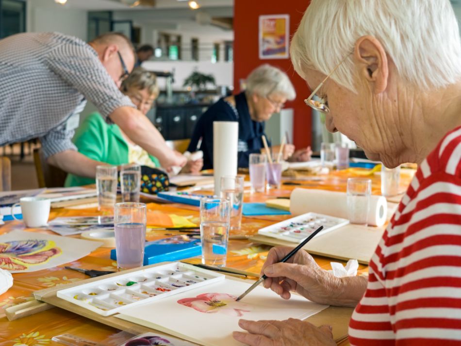 Eine Gruppe älterer Menschen sitzt an einem Tisch und malt mit Wasserfarben. 