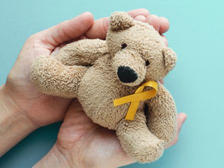 Hände halten einen Teddy mit einer gelb-goldenen Awareness-Schleife.