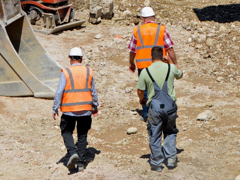Rückansicht von drei Bauarbeitern, die in der Sonne über eine Baustelle laufen.