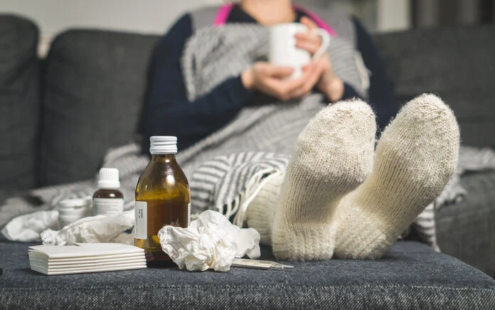 Erkältung und Fieber, © Tero Vesalainen, Shutterstock Im Bildvordergrund sind Medikamente und Taschentücher zu sehen; im Bildhintergrund liegt eine zugedeckte Frau auf einem Sofa, die eine Tasse mit beiden Händen hält.
