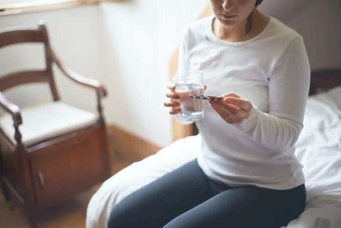 Junge Frau sitzt auf dem Bett mit einem Tablettenblister in der einen und einem Glas Wasser in der anderen Hand.