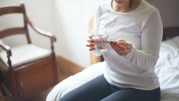Tabletteneinnahme, © Kampus Production, Pexels Junge Frau sitzt auf dem Bett mit einem Tablettenblister in der einen und einem Glas Wasser in der anderen Hand.