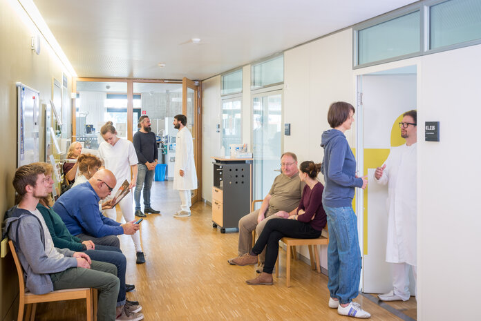 Wartezimmer, © Krebsinformationsdienst, DKFZ; Foto: Tobias Schwerdt Mehrere Patienten und Patientinnen sitzen im Wartezimmer einer Tagesklinik oder Chemo-Ambulanz. Ärzte stehen im Flur und in der Tür zum Behandlungszimmer.