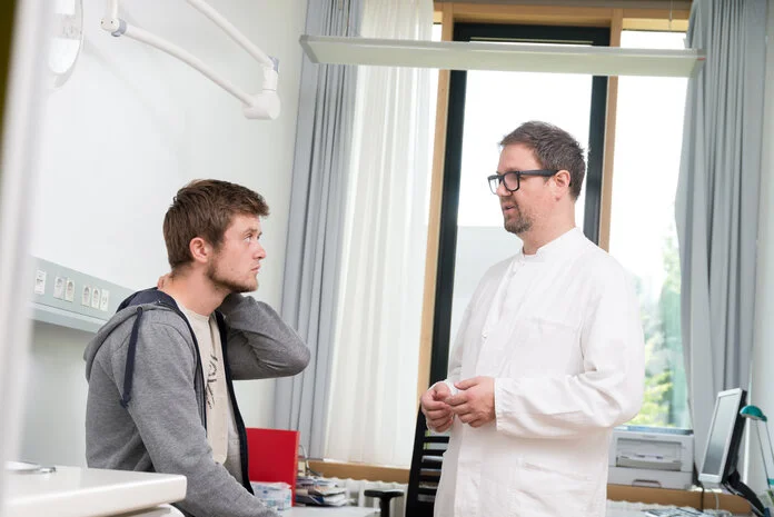 Aufklärungsgespräch, © Krebsinformationsdienst, DKFZ; Foto: Tobias Schwerdt Ein junger Mann sitzt auf der Untersuchungsliege. Vor ihm steht der Arzt.