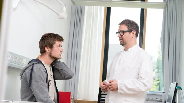 Aufklärungsgespräch, © Krebsinformationsdienst, DKFZ; Foto: Tobias Schwerdt Ein junger Mann sitzt auf der Untersuchungsliege. Vor ihm steht der Arzt.