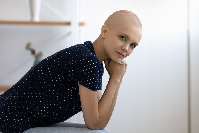 Eine junge Patientin mit Glatze sitzt auf einer Bettkante und denkt nach.