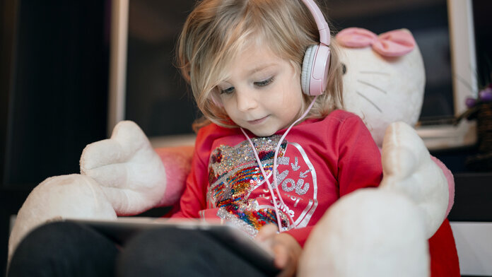 Kleines Kind sitzt auf einem Kindersofa, trägt Kopfhörer und schaut auf ein Tablet.