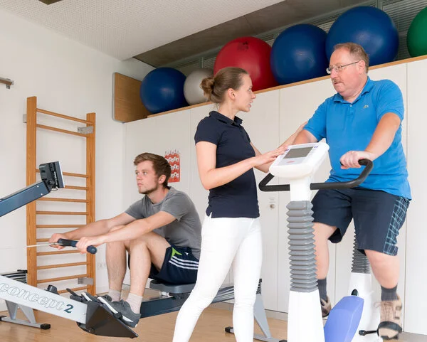 Physiotherapie Fahrradergometer, © Krebsinformationsdienst, DKFZ; Foto: Tobias Schwerdt Ein Mann fährt in einem Sportraum von einer Physiotherapeutin begleitet auf dem Fahrradergometer. Im Hintergrund trainiert ein junger Mann auf einem Rudergerät.
