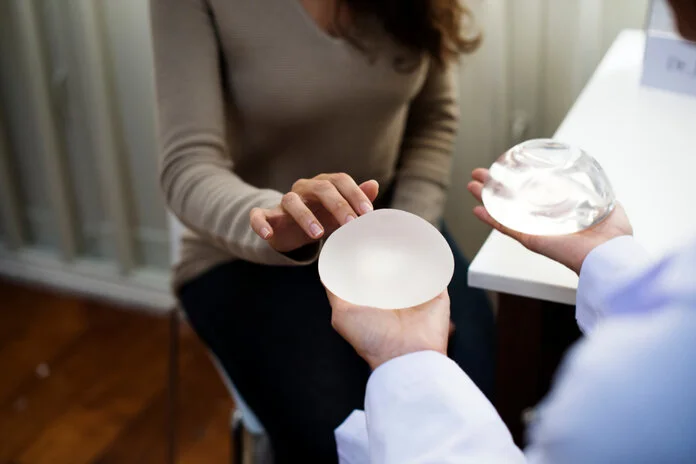 Brustimplantate mit glatter und aufgerauter Oberfläche, Rawpixel.com/shutterstock Arzt hält ein Brustimplantat mit glatter und ein Implantat mit aufgerauter Oberfläche in der Hand, während eine Frau das linke Implantat berührt. © Rawpixel.com, Shutterstock
