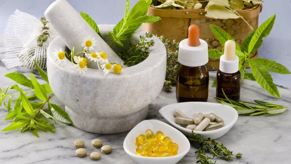 Komplementäre und alternative Methoden, © Pat_Hastings, Shutterstock Mörser mit Kräutern, Blüten und Pistill auf Marmorplatte mit weiteren Kräutern, Blättern in Holzkorb, Schälchen mit Dragees und Tabletten sowie zwei Braunglasflaschen mit Pipette.