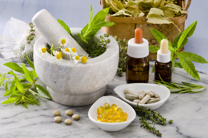 Komplementäre und alternative Methoden, © Pat_Hastings, Shutterstock Mörser mit Kräutern, Blüten und Pistill auf Marmorplatte mit weiteren Kräutern, Blättern in Holzkorb, Schälchen mit Dragees und Tabletten sowie zwei Braunglasflaschen mit Pipette.