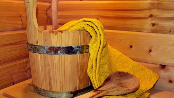 Sauna, © ulleo, Pixabay Wassereimer aus Holz, Holzschöpflöffel und gelbes Handtuch in einer Holzsauna.