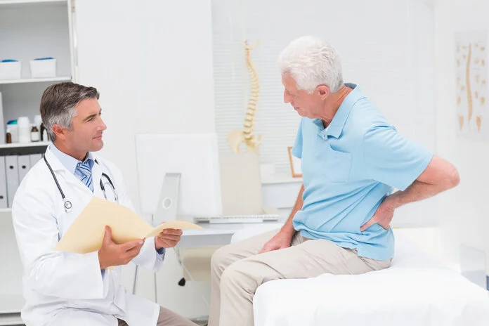 Arztgespräch zu Rückenschmerzen, © ESB Professional, Shutterstock Ein Arzt sitzt mit einer aufgeklappten Patientenakte vor einem älteren Patienten, der auf einer Patientenliege sitzt und sich den Rücken hält.