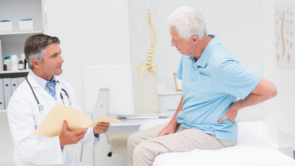 Ein Arzt sitzt mit einer aufgeklappten Patientenakte vor einem älteren Patienten, der auf einer Patientenliege sitzt und sich den Rücken hält.