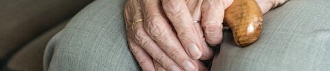 Nahaufnahme einer Seniorin, die mit übereinander gelegten Händen auf dem Sofa sitzt.