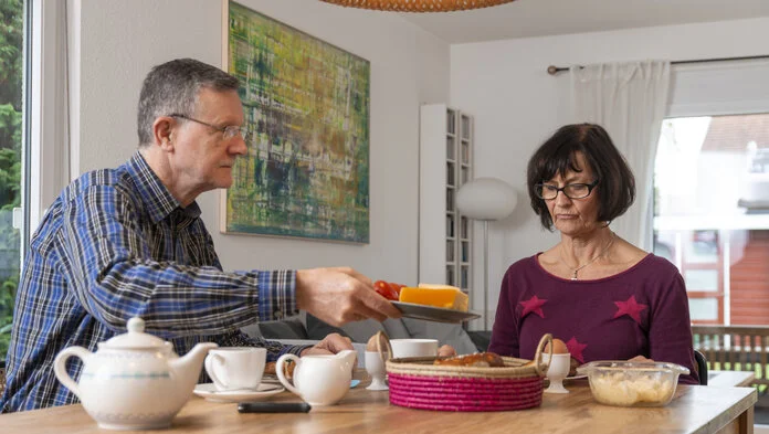 Ein Mann bietet einer Frau am Frühstückstisch etwas zu essen an.