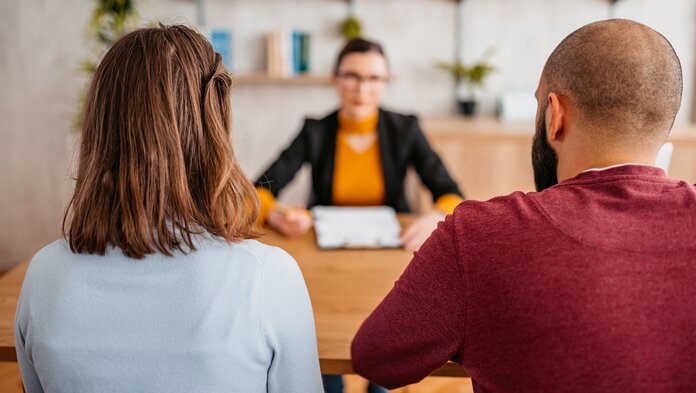 Paar sitzt mit Rücken zum Betrachter an einem Tisch einer Frau gegenüber, vor der Papierunterlagen liegen.