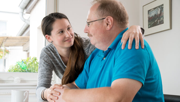 Junge Frau lächelt älteren Mann an, während sie ihn mit einem Arm umarmt und ihre andere Hand auf seine Hand legt.