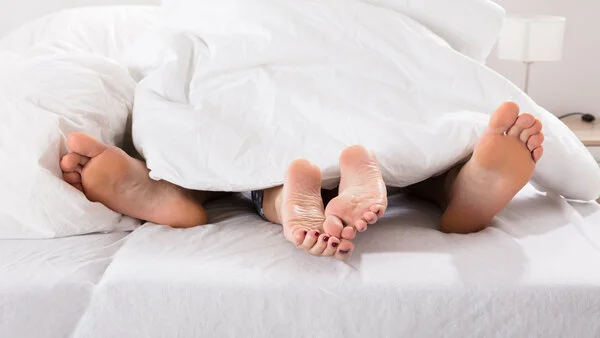 Ein Paar im Bett unter der Bettdecke. Zu sehen sind ihre Füße.