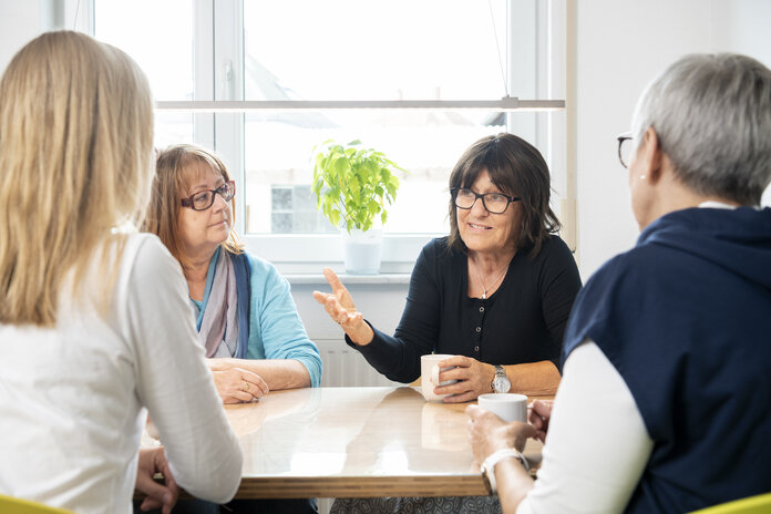 4 Frauen mittleren Alters sitzen zusammen an einem Tisch, trinken Kaffee und reden miteinander. Eine Frau redet, während die anderen 3 zuhören.