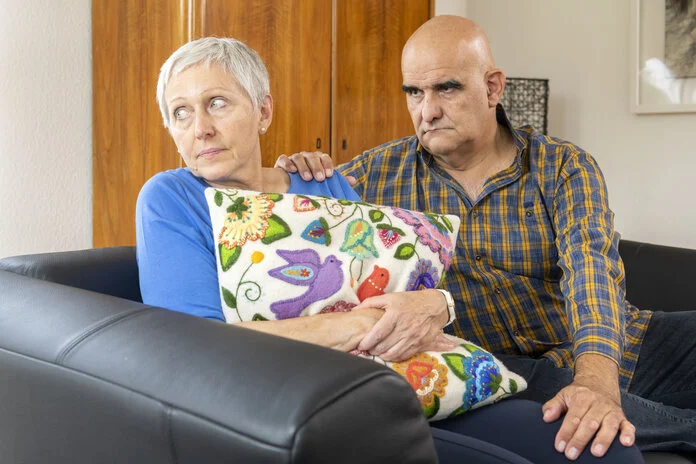 Mann tröstet Frau, © Krebsinformationsdienst, DKFZ; Foto: Tobias Schwerdt Ein älteres Paar sitzt auf dem Sofa. Die Frau hält ein Kissen an sich gedrückt und schaut traurig weg. Der Mann hat tröstend die Hand auf die Schulter der Frau gelegt und schaut betreten.