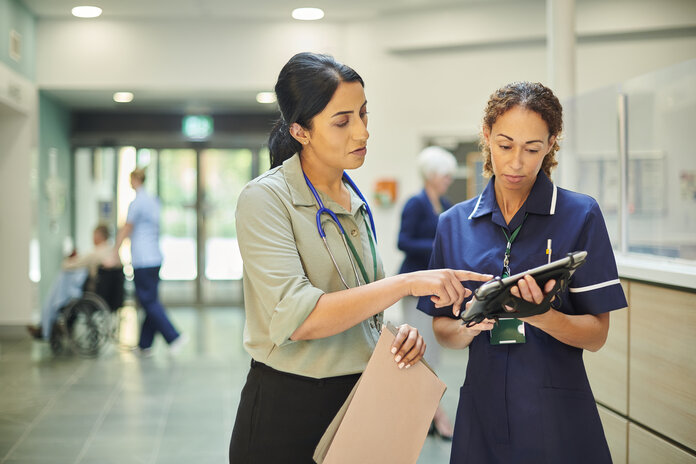 Patientenakte in Papierform oder digital , © sturti, iStock Eine Ärztin und eine Pflegerin stehen im Klinikflur und schauen gemeinsam auf ein Tablet. Die Ärztin hält ausgedruckte Dokumente in einer Hand.