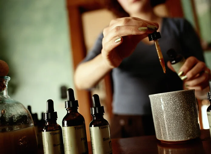 Homöopathische Tropfen Eine Frau hält eine Pipettenflasche in der Hand und tropft etwas in eine Tasse, im Vordergrund stehen weitere braune Pipettenflaschen.