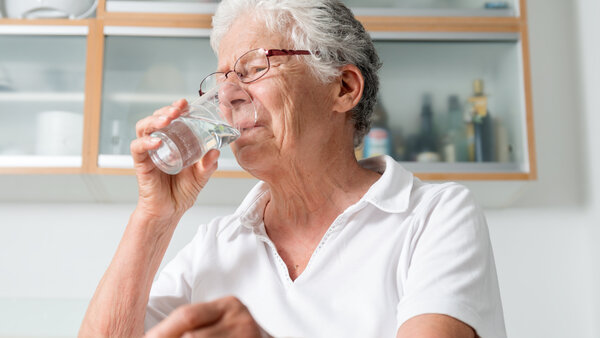 Ältere Frau sitzt in der Küche und trinkt Wasser aus einem Glas.