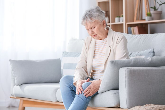 Gelenkschmerzen im Knie , © Pixel-Shot, Shutterstock Ältere Frau sitzt auf dem Sofa und umfasst schmerzerfüllt ihr Knie.