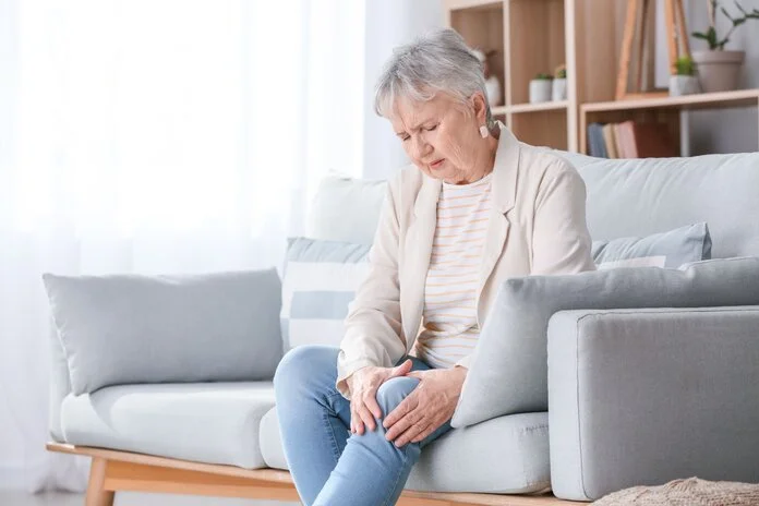 Gelenkschmerzen im Knie , © Pixel-Shot, Shutterstock Ältere Frau sitzt auf dem Sofa und umfasst schmerzerfüllt ihr Knie.