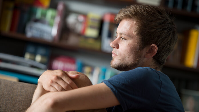 Junger Mann auf der Couch, der seine Arme um die Beine geschlungen hat und nachdenklich schaut.