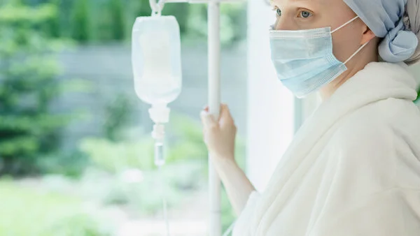 Patientin mit Mundschutz, © Ground Picture, Shutterstock Patientin mit Kopftuch und Mundschutz hält einen Infusionsständer und schaut aus dem Fenster.