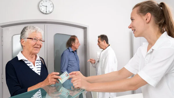 Arztpraxis: Empfang, © Krebsinformationsdienst, DKFZ; Foto: Tobias Schwerdt Patientin reicht einer Arzthelferin ihre Versichertenkarte am Empfang einer Arztpraxis. Im Hintergrund dpricht ein Arzt mit einem Patienten.