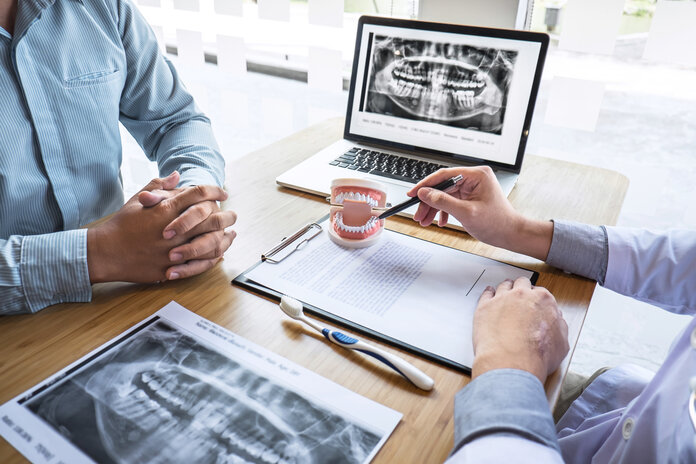 Beratung Zahnpflege, © SuperOhMo, Shutterstock Die Hände eines Zahnarztes deuten auf ein Gebiss, daneben liegen eine Zahnbürste und Röntgenbilder der Zähne. Ein Mann sitzt gegenüber.