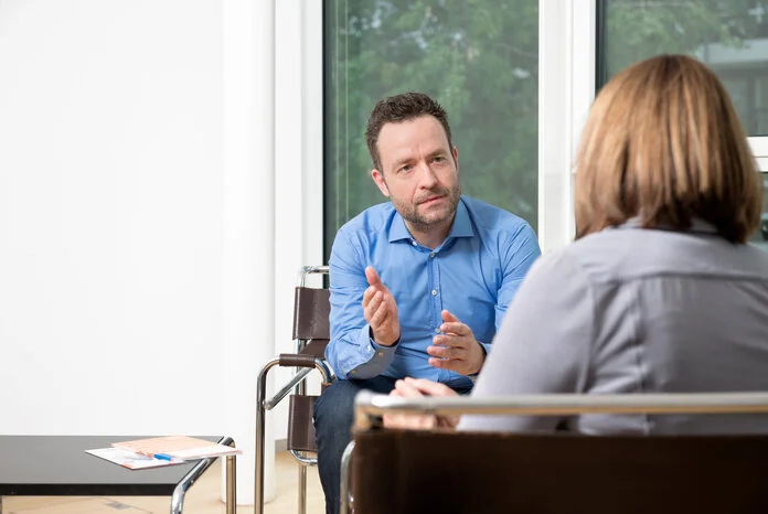 Eine Frau, von hinten fotografiert, sitzt einem Mann im Beratungsgespräch gegenüber.
